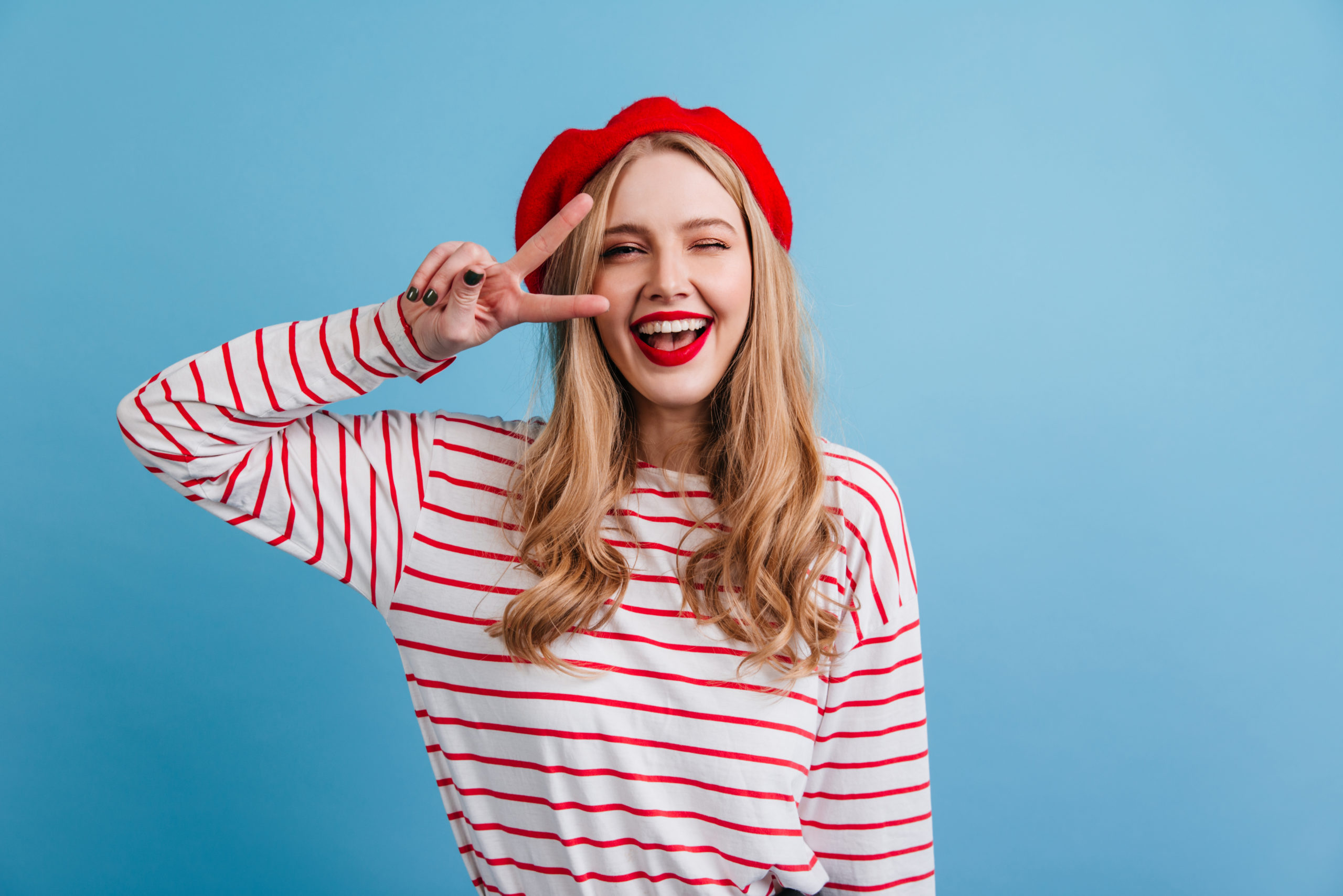 Pretty French blonde girl in striped shirt showing peace sign. Front view of laughing french lady posing on blue background..